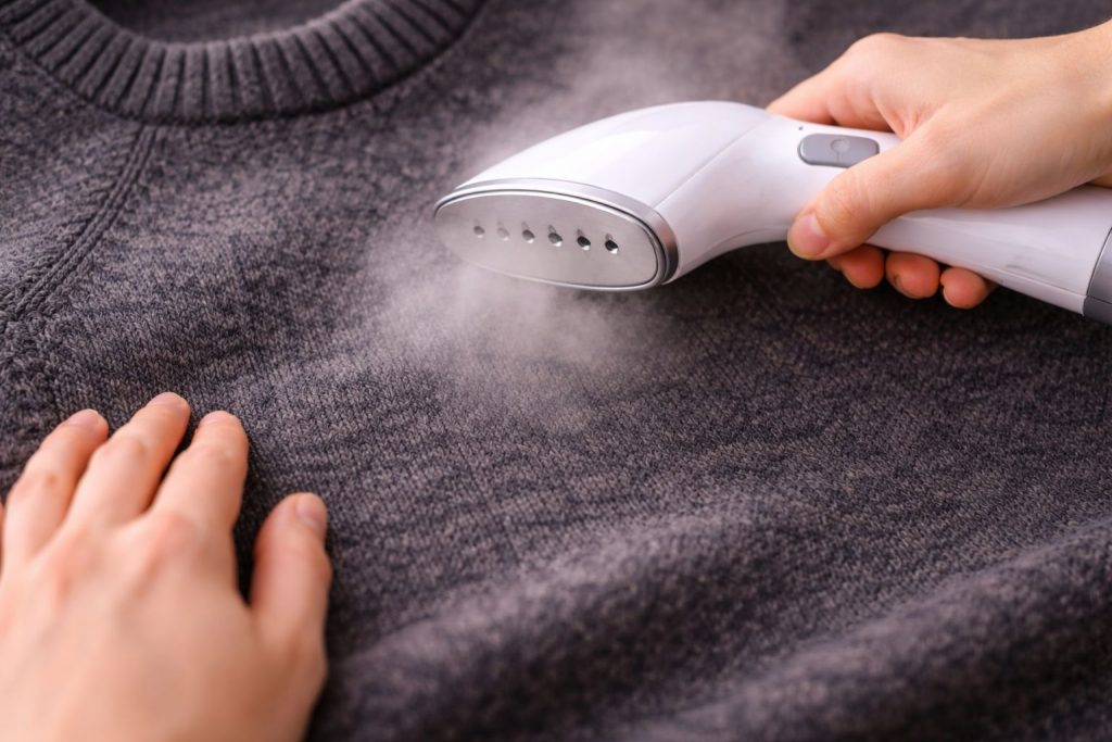 Close-up on hands carefully steaming a dark grey merino wool sweater, capturing the texture of the knit and the visible steam. Soft, warm lighting.