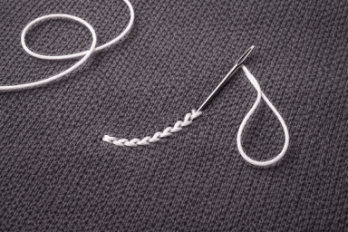Minimalist top-down photo, an embroidery needle with smooth white thread stitching onto dark gray knit fabric. Centered composition, clean studio lighting.