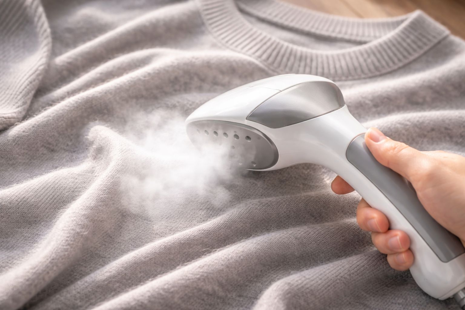 A close-up of a handheld steamer releasing steam onto a wrinkled merino wool sweater