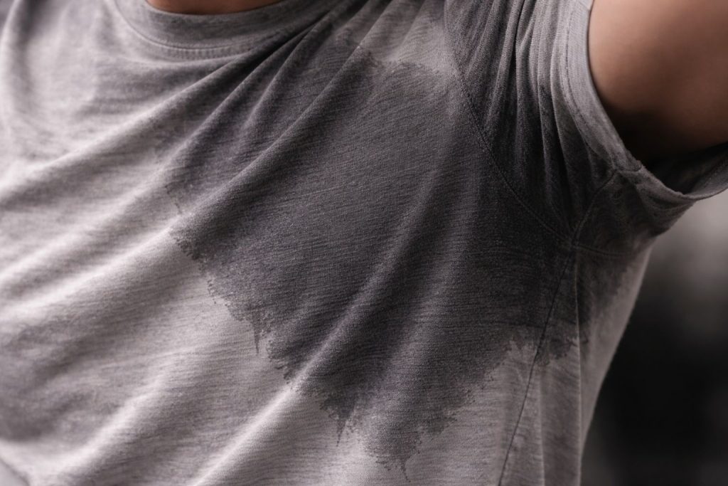 A powerful close-up of a dark, wet sweat stain saturating a grey cotton t-shirt. The texture should look heavy and damp, clinging to the skin. Photorealistic style.
