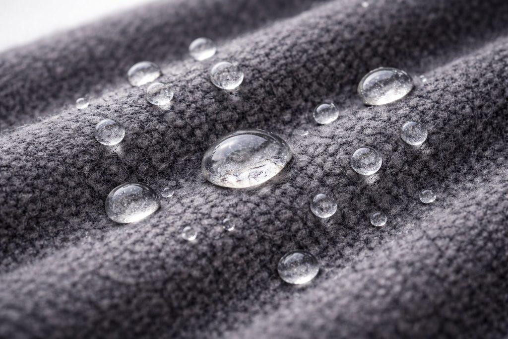 Water droplets bead on the surface of a dark grey merino wool fabric, clearly showcasing its water-resistance. Soft light, clean background.