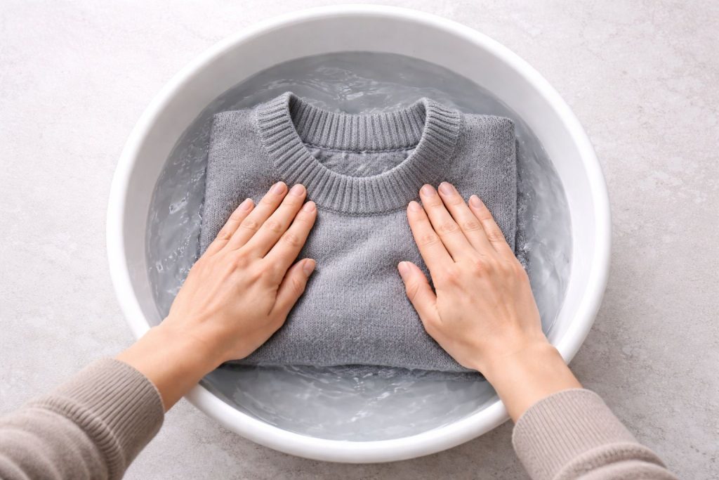 A minimalist top-down shot of a person's hands gently pressing water out of a neatly folded, high-quality gray merino wool sweater in a clean white basin. The water is clear, with soft, natural l